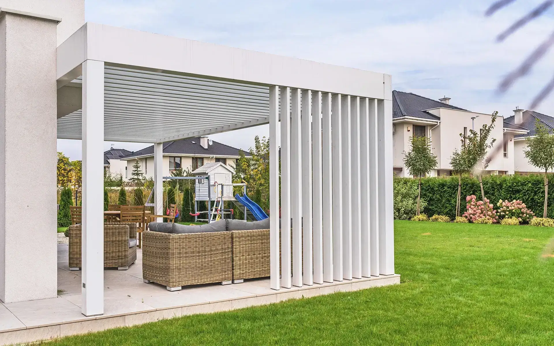 Auf dem Bild sieht man eine moderne weiße Pergola neben einem Einfamilienhaus. Unter der Überdachung stehen Gartenmöbel, und im Hintergrund befindet sich ein großer Garten mit Rasen sowie ein Spielplatz für Kinder.