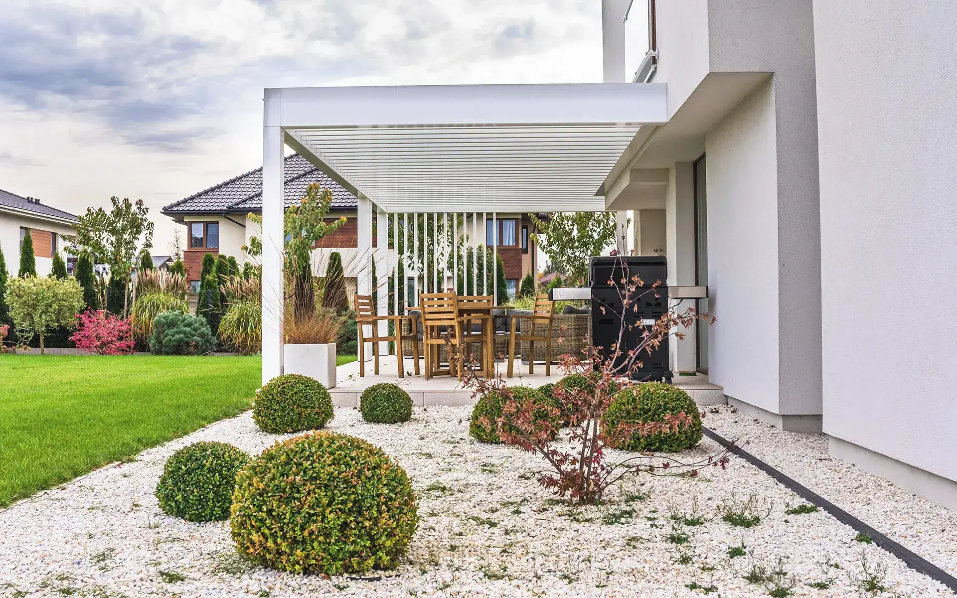 Auf dem Bild sieht man eine moderne Terrasse mit einer weißen Pergola neben einem Einfamilienhaus. Unter der Überdachung stehen ein Tisch mit Stühlen und ein Grill, umgeben von einem gepflegten Garten mit Rasen und Zierpflanzen.