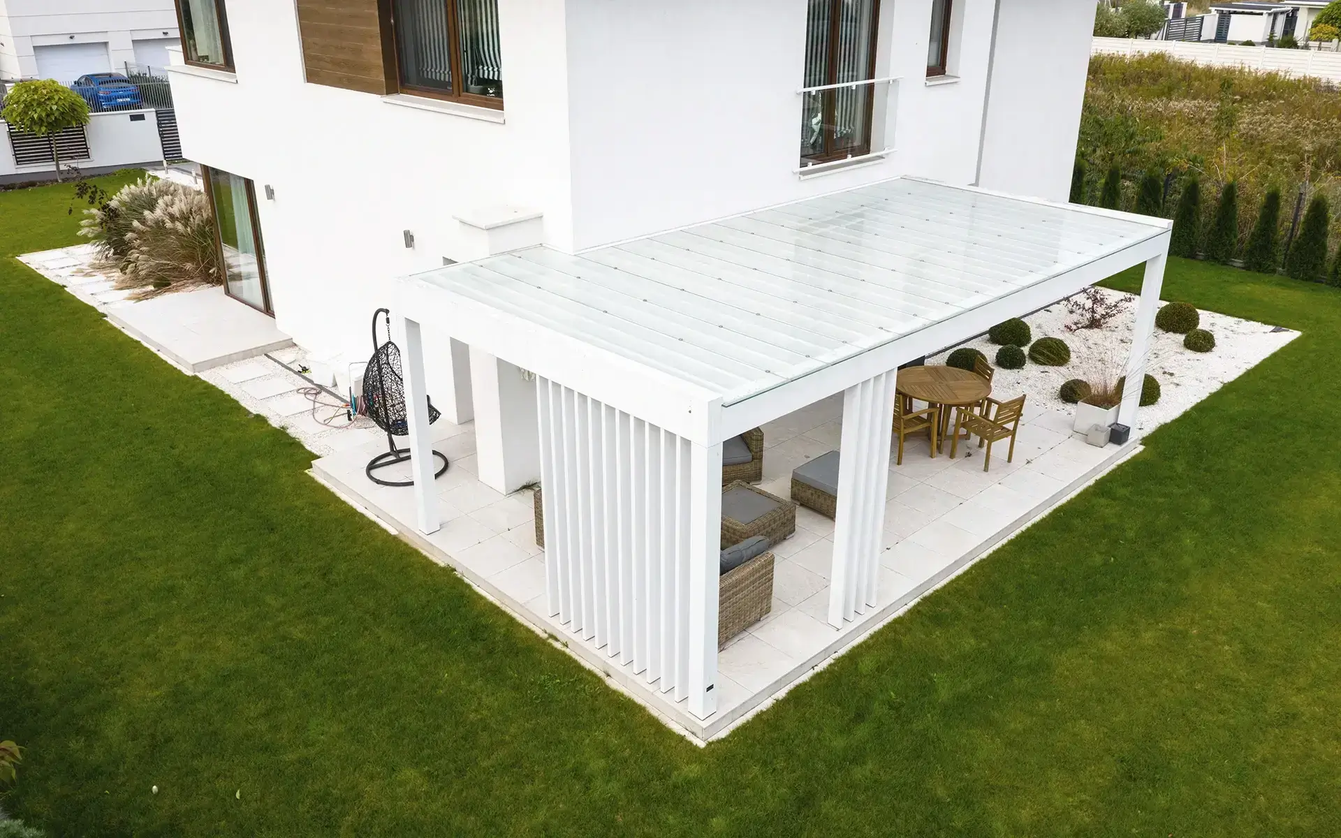 Auf dem Bild sieht man eine moderne weiße Pergola neben einem Einfamilienhaus, aufgenommen aus der Vogelperspektive. Unter der Überdachung befinden sich Gartenmöbel und ein Essbereich, umgeben von einem gepflegten Garten mit Rasen und Zierpflanzen.