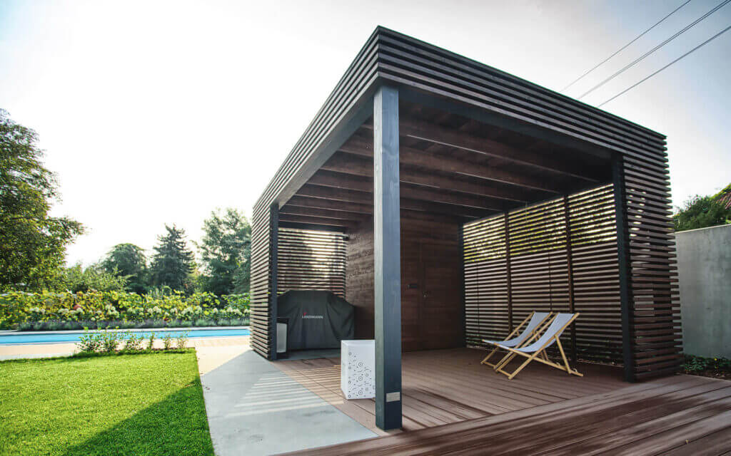 Auf dem Bild sieht man eine moderne Holzpergola im Garten neben einem Pool. Darunter stehen zwei Liegestühle und ein Grill, während Sonnenlicht durch die Lamellen fällt.