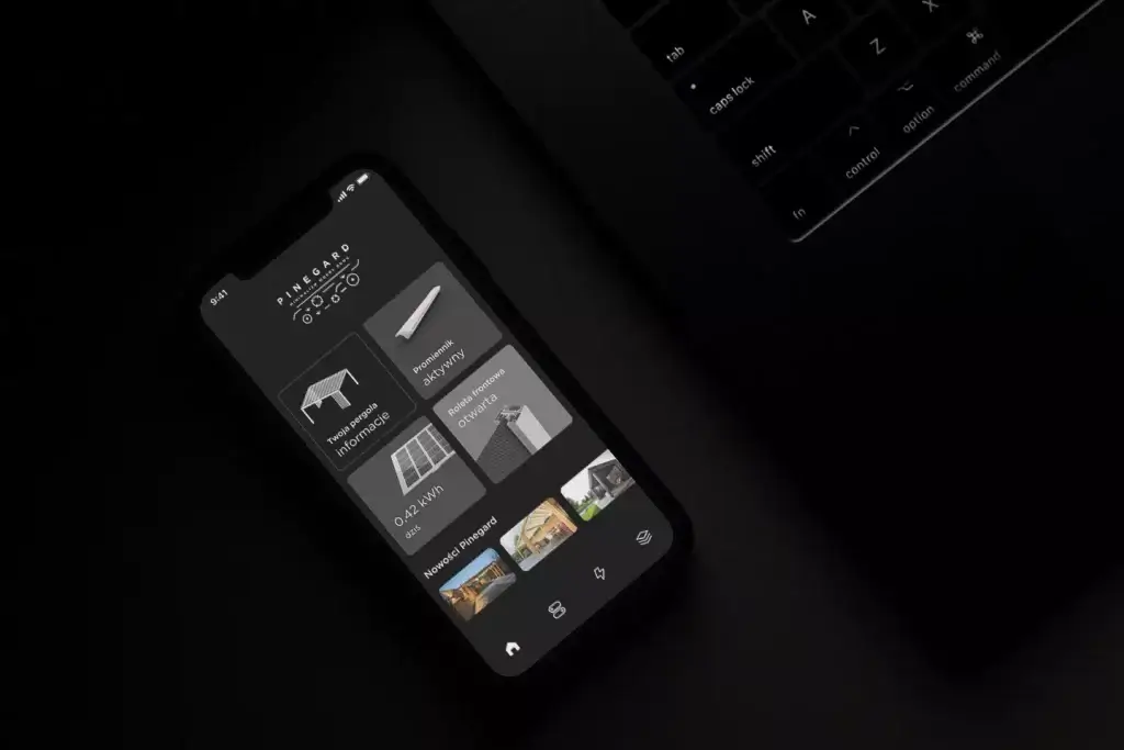 Auf dem Bild sieht man ein Smartphone auf einem dunklen Tisch neben einer Laptop-Tastatur. Auf dem Display ist eine App von PINEGARD zu sehen, mit der man eine Pergola und ihre Funktionen steuern kann.