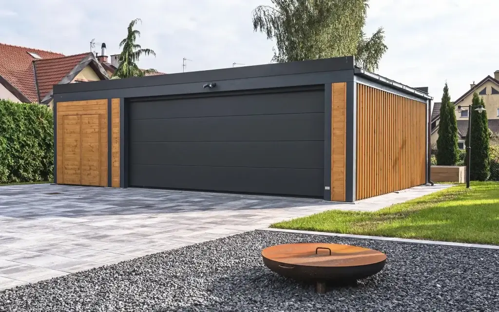 Carport garażowy na zdjęciu to nowoczesna, wolnostojąca konstrukcja o minimalistycznym designie. Dominują ciemne, stonowane kolory — grafitowe drzwi garażowe zestawione z pionowymi panelami z naturalnego drewna. Płaski dach podkreśla geometryczną formę budynku, a otoczenie — starannie ułożona kostka, zielony trawnik i dekoracyjny żwir — nadaje całości uporządkowany, elegancki charakter. Na pierwszym planie znajduje się metalowe palenisko, które dodaje przestrzeni funkcjonalności i przytulności.
