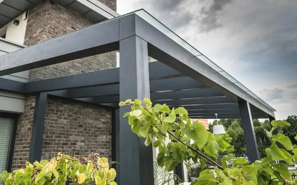 Auf dem Bild sieht man eine moderne Pergola mit dunklen Metallstützen neben einem Haus aus Backstein. Im Vordergrund befinden sich grüne Pflanzen, und darüber ist ein bewölkter Himmel zu sehen.