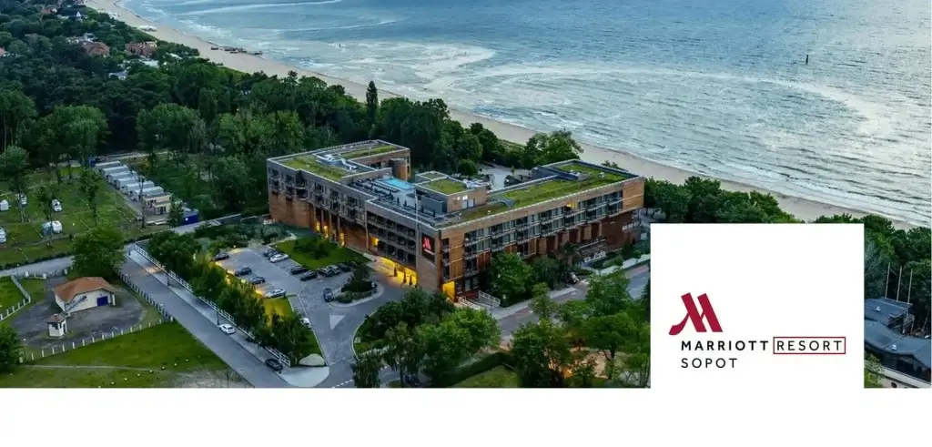 Zdjęcie przedstawia widok z lotu ptaka na Marriott Resort w Sopocie, położony tuż przy plaży, z widoczną linią brzegową i falami w tle. Budynek ma nowoczesną bryłę z płaskim dachem, otoczony zielenią i drzewami, co nadaje mu spokojny, nadmorski charakter. Na terenie wokół hotelu widać zaparkowane samochody oraz uporządkowaną przestrzeń, sugerującą wygodny dostęp dla gości. Całość emanuje elegancją i harmonią z otoczeniem, idealnie wpisując się w krajobraz kurortu. Logo „Marriott Sopot Resort” w prawym dolnym rogu podkreśla prestiż i lokalizację obiektu, zachęcając do wypoczynku w luksusowych warunkach nad Bałtykiem.