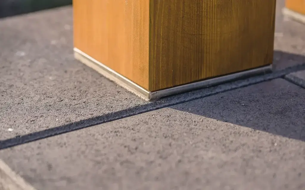 Das Foto zeigt den unteren Teil eines Holzpfostens, der auf einer Oberfläche aus Beton- oder Steinplatten befestigt ist. Am Sockel des Pfostens ist eine Metallleiste angebracht, die der Konstruktion ein ästhetisches und langlebiges Aussehen verleiht.