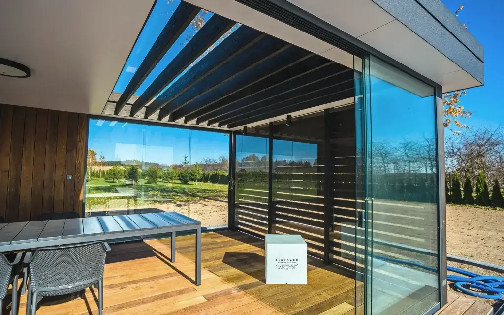 Auf dem Bild sieht man eine moderne Terrasse mit Glaswänden und einer Pergola mit dunklen Lamellen. Drinnen stehen ein Tisch mit Stühlen und ein weißer Würfel mit dem Logo „PINEGARD“.