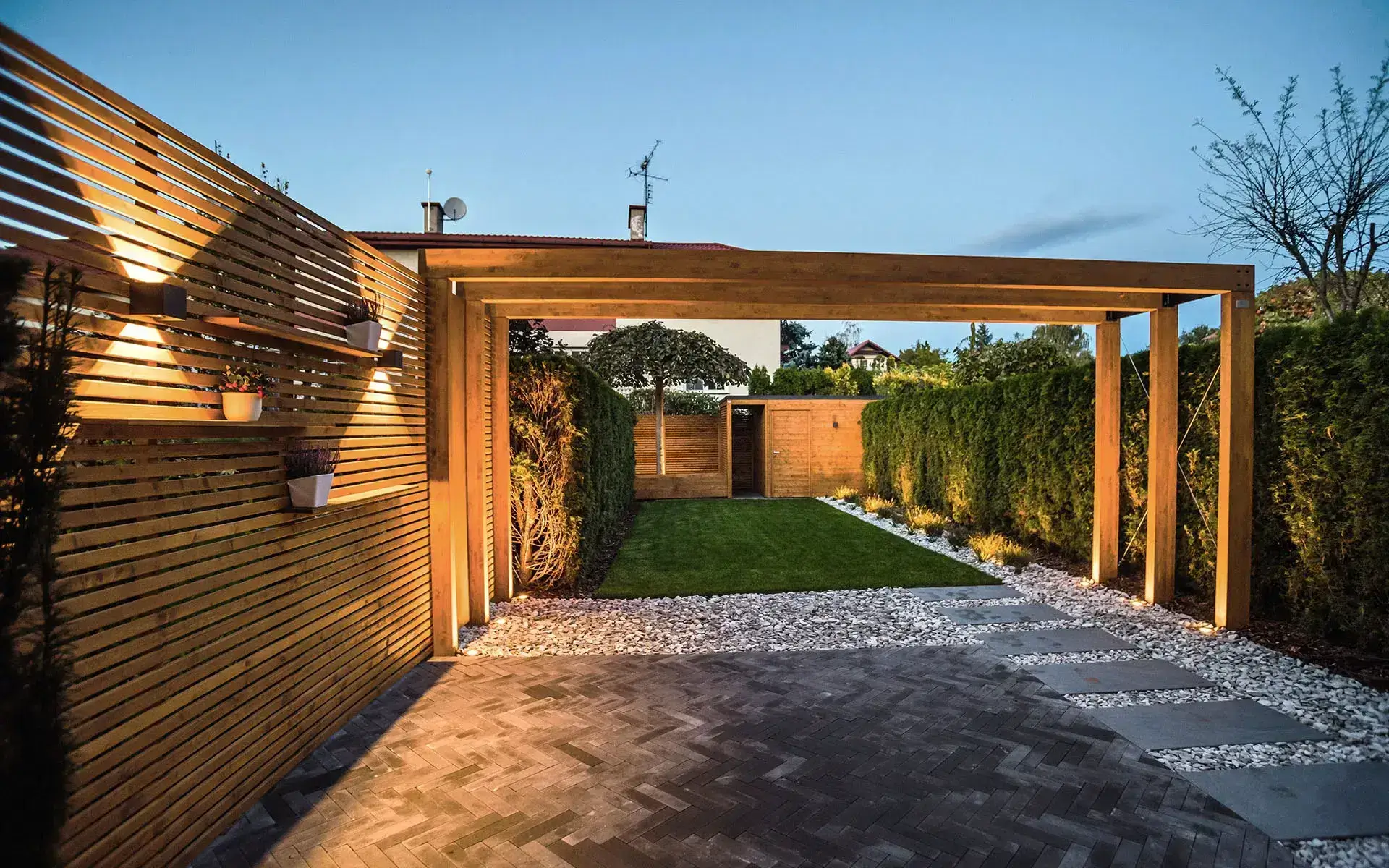 Stylowa pergola ogrodowa z płaskim dachem i pionowymi lamelami tworzy przytulną strefę wypoczynku wśród zieleni. Ciepłe oświetlenie, donice z roślinami i ściana z dekoracyjnymi skrzynkami nadają przestrzeni kameralny, nowoczesny charakter. Posadzka z cegły w jodełkę płynnie przechodzi w żwirową ścieżkę z płytami prowadzącą do trawnika. Całość harmonijnie łączy funkcjonalność, estetykę i naturalne otoczenie.