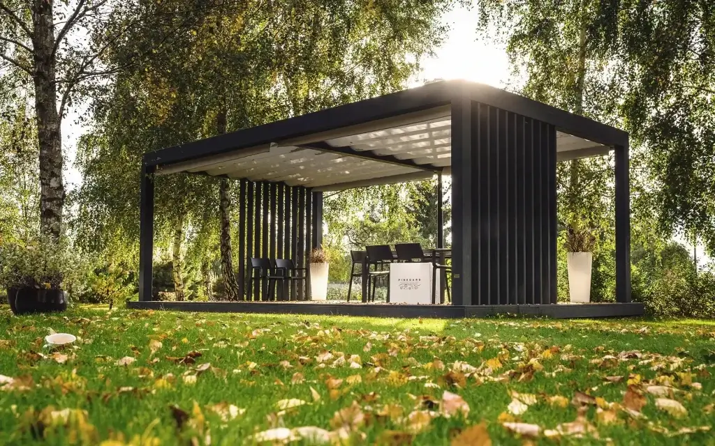 Nowoczesna pergola ogrodowa z płaskim dachem i pionowymi lamelami — elegancka przestrzeń do wspólnych posiłków w otoczeniu drzew. Wewnątrz stół z krzesłami, a narożne donice z zielenią dodają przytulności. Jesienne liście na trawie i miękkie światło tworzą ciepłą, naturalną atmosferę. Idealne miejsce na relaks w harmonii z przyrodą.
