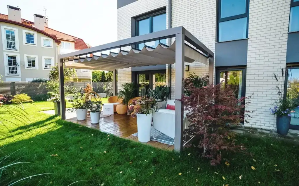 Auf dem Bild sieht man eine Terrasse mit einer modernen Pergola und einem hellen Stoffdach. Darunter stehen Gartenmöbel und mehrere große Blumentöpfe mit Pflanzen.
