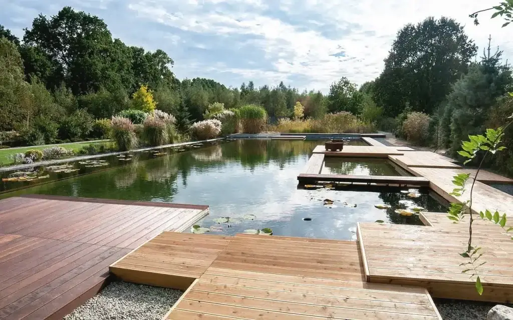 Auf dem Bild sieht man einen großen Naturpool, der von Holzstegen umgeben ist. Rundherum wachsen viele Pflanzen und Bäume, die sich im ruhigen Wasser spiegeln.
