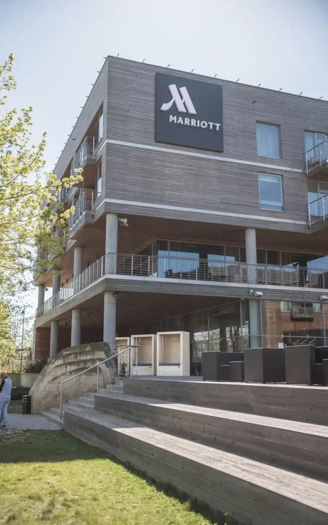 Na zdjęciu widoczny jest nowoczesny, wielokondygnacyjny budynek hotelowy z wyraźnym logo Marriott, co wskazuje na jego przynależność do znanej sieci. Fasada łączy drewniane panele z dużymi przeszkleniami i balkonami, tworząc elegancką, współczesną kompozycję. Przed hotelem znajduje się zagospodarowana strefa wypoczynkowa z drewnianymi schodami i miejscami do siedzenia, otoczona zielenią. Drzewa i roślinność nadają przestrzeni przyjazny, relaksujący charakter, zachęcający gości do spędzania czasu na świeżym powietrzu.