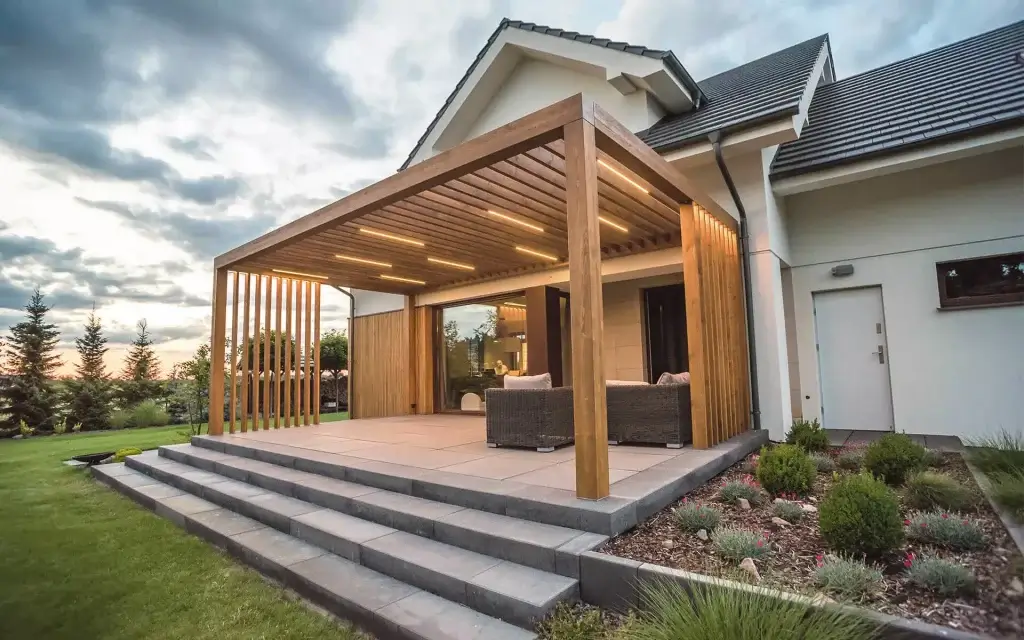 Na zdjęciu widać nowoczesny taras przy domu jednorodzinnym z drewnianą pergolą. Pergola ma prostą, geometryczną konstrukcję z jasnego drewna i płaski dach wykonany z równoległych listew. W suficie zamontowane są podłużne lampy LED, które oświetlają przestrzeń pod zadaszeniem i tworzą przytulny klimat.
