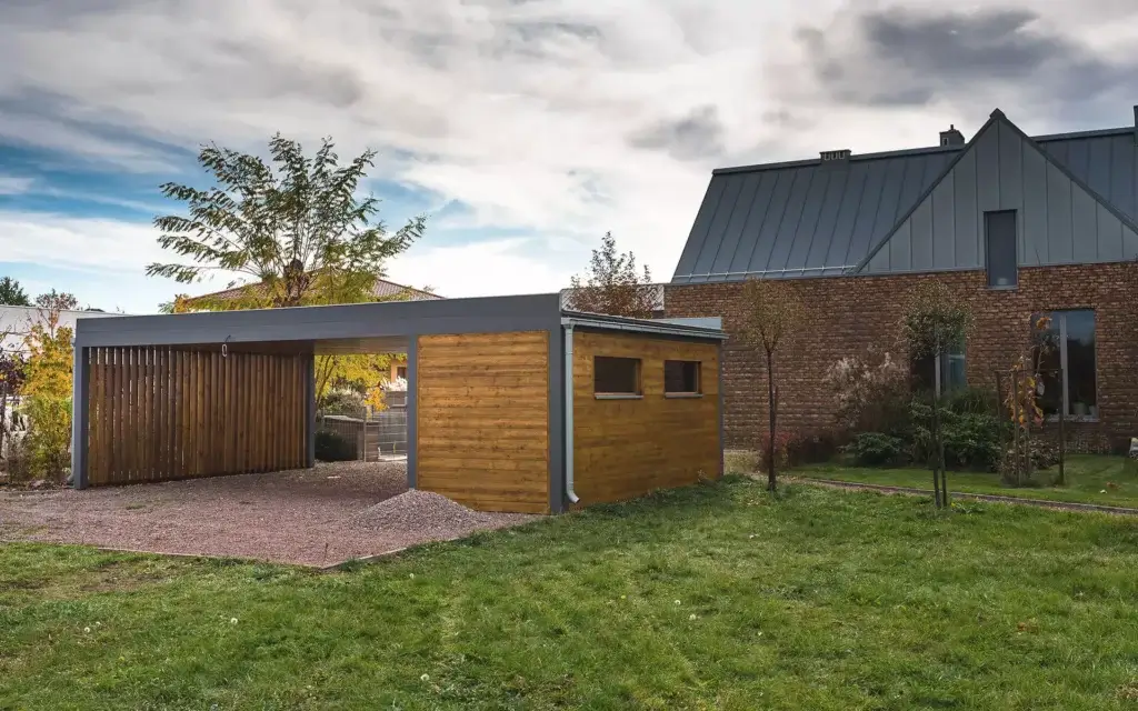 Na zdjęciu znajduje się nowoczesny carport z drewna, zintegrowana z zamkniętym schowkiem po jednej stronie. Obok stoi współczesny dom o ceglanej elewacji i metalowym dachu, otoczony zadbaną zielenią.