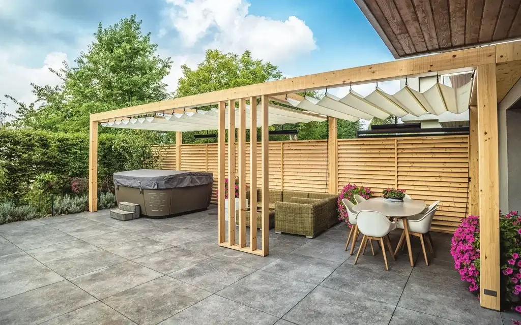Das Foto zeigt einen Ausschnitt einer modernen Gartenkonstruktion aus Holz. Im Zentrum befindet sich ein Paneel mit horizontalen Latten aus hellem Holz, eingebettet in einen dunkleren Rahmen. Die Latten sind leicht geneigt, was eine Belüftungs- oder Dekorationsfunktion haben könnte. Der Hintergrund ist verschwommen und zeigt Grünpflanzen und Vegetation.