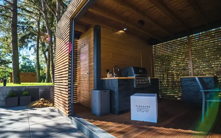 Auf dem Bild sieht man eine moderne Gartenküche aus Holz in einem überdachten Pavillon. Im Inneren befinden sich ein Grill, eine Arbeitsplatte mit Spüle sowie Sitzmöbel, umgeben von einem grünen Garten mit Bäumen.