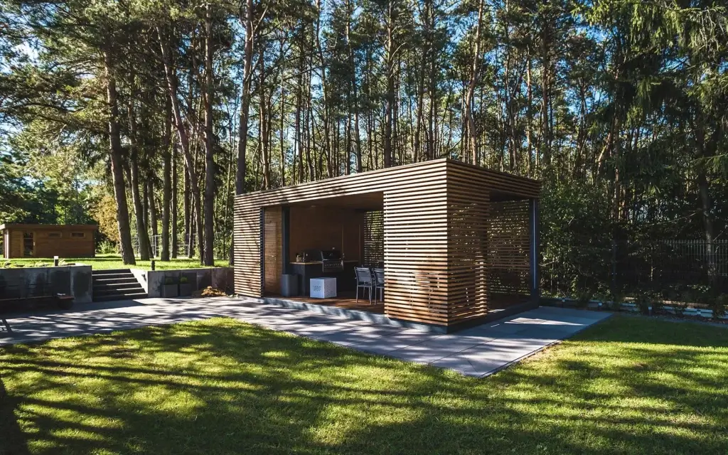 Nowoczesna altana z drewnianych lameli wśród wysokich sosen — półotwarta, elegancka, idealna do wypoczynku i spotkań. W środku stół z krzesłami, wokół zadbany trawnik, kamienne stopnie i spójna architektura w tle. Naturalna harmonia i funkcjonalność.