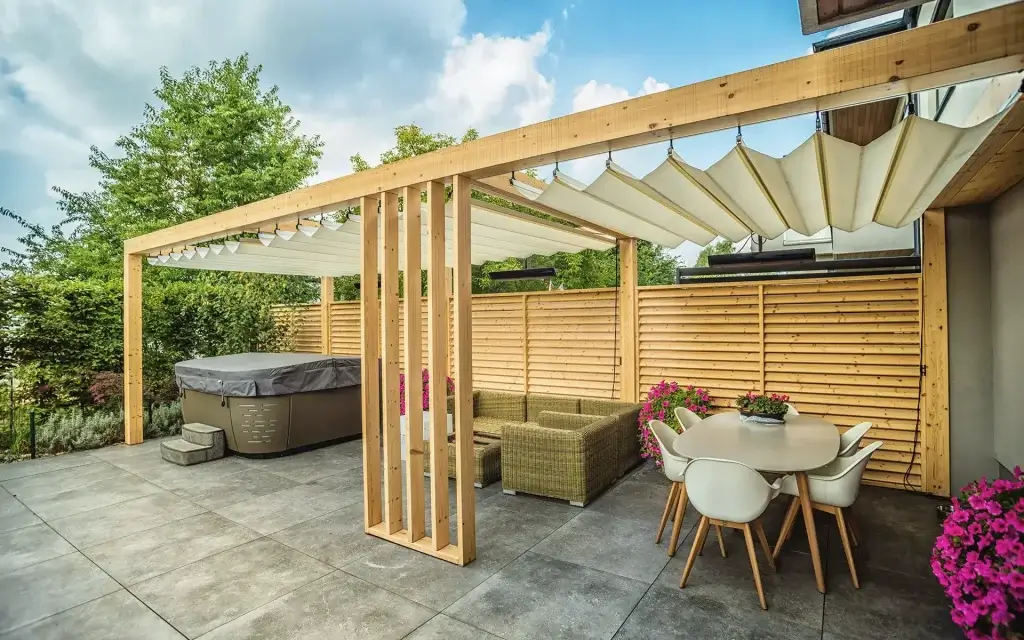 Na zdjęciu widać przestronny taras z ogrodową konstrukcją pergolową, tworzącą salon wypoczynkowy ze strefą spa. Pergola wykonana jest z jasnego drewna i ma prostą, nowoczesną konstrukcję z poziomą belką nośną. Dach tworzy rozkładana markiza materiałowa, która zapewnia cień i ochronę przed słońcem. Po jednej stronie pergoli znajduje się ścianka z pionowych drewnianych lameli.
