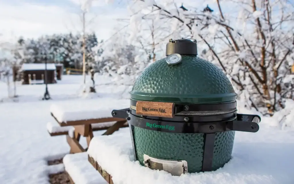 Na zdjęciu widać zielony grill ceramiczny Big Green Egg ustawiony na zewnątrz w zimowym ogrodzie, częściowo otoczony śniegiem.