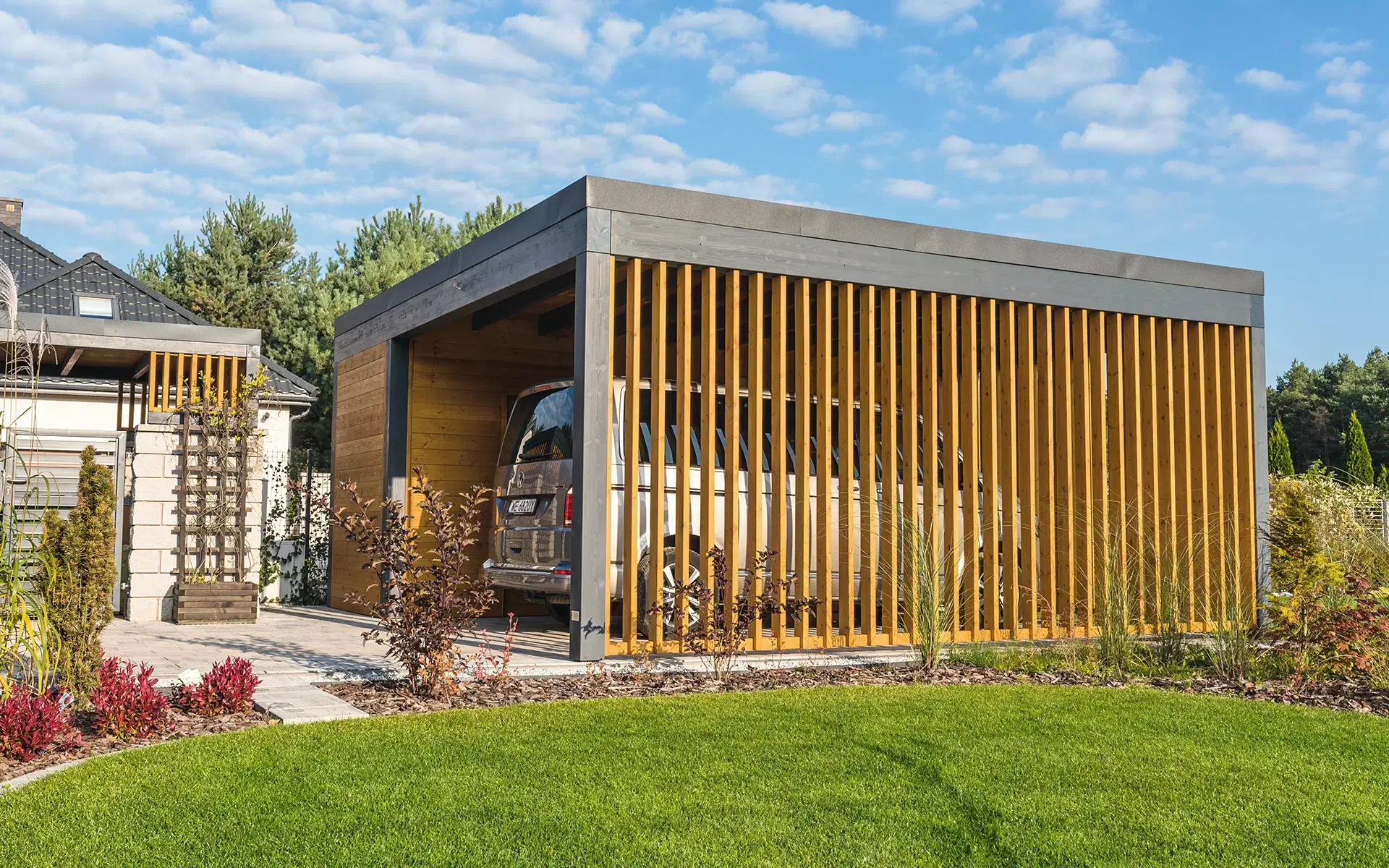 Nowoczesny carport z pionowymi drewnianymi lamelami i płaskim dachem — elegancka osłona dla auta w otoczeniu zadbanego ogrodu.