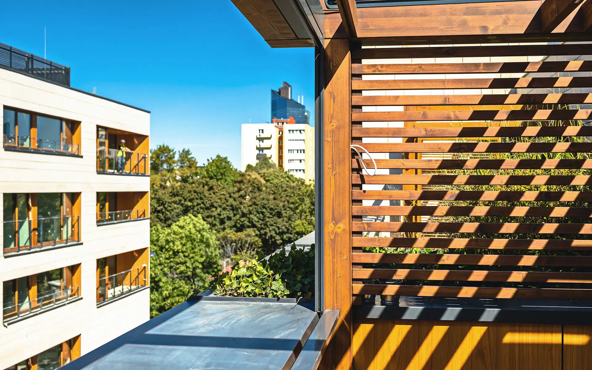 Zdjęcie przedstawia nowoczesny balkon z widokiem na miejską zabudowę, gdzie natura subtelnie przeplata się z architekturą. W pierwszym planie widoczna jest drewniana osłona z poziomymi listwami, rzucająca dekoracyjne cienie na ścianę i podłogę. Balkon wyposażony jest w metalową balustradę oraz niewielki ogródek z zielonymi roślinami, dodającymi świeżości. W tle rozciągają się wielopiętrowe budynki mieszkalne z dużymi oknami i balkonami, otoczone drzewami. Na horyzoncie widać nowoczesny biurowiec, a całość tonie w jasnym, błękitnym świetle dnia, tworząc obraz komfortowego życia w mieście.