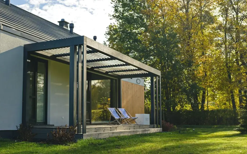 Nowoczesna altana ogrodowa z czarną pergolą i drewnianym tarasem — minimalistyczna, elegancka, otwarta na zieleń. Dwa leżaki ustawione w stronę słońca zachęcają do relaksu, a duże przeszklenia domu płynnie łączą wnętrze z naturą. Przestrzeń pełna światła i spokoju.