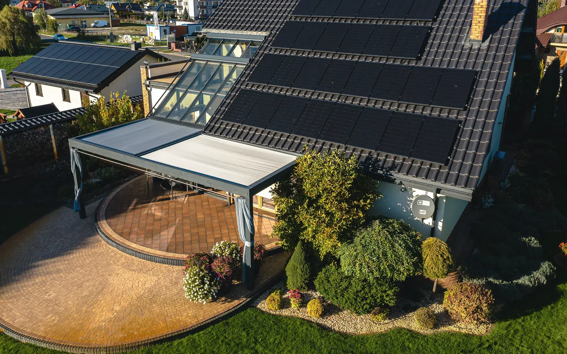Auf dem Bild sieht man eine Luftaufnahme eines Hauses mit großem Dach, das mit mehreren Solarpanelen ausgestattet ist. Darunter befindet sich eine elegante Pergola mit Stoffdach über einer runden Terrasse, umgeben von dekorativen Sträuchern und Blumenbeeten.