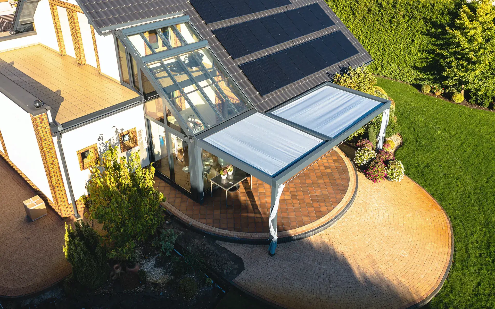 Auf dem Bild sieht man eine Luftaufnahme eines Hauses mit moderner Glasveranda und einer angrenzenden Pergola mit hellem Stoffdach. Auf dem Dach sind mehrere Solarpaneele installiert, und die Terrasse ist von gepflegtem Rasen sowie dekorativen Pflanzen umgeben.