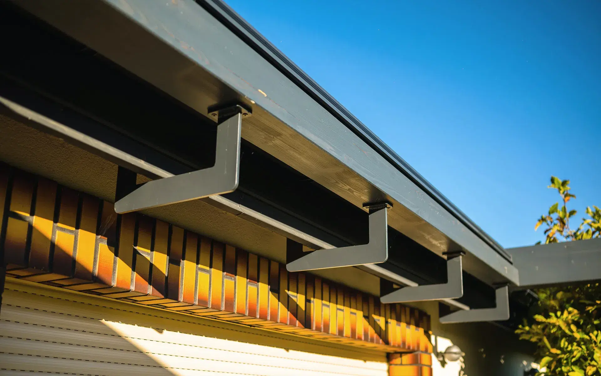 Auf dem Bild sieht man eine Detailaufnahme der modernen Pergola-Konstruktion mit stabilen, dunkelgrauen Metallhalterungen unter dem Dach. Darunter befindet sich eine Hausfassade aus Ziegeln und ein Rollladen, beleuchtet vom warmen Sonnenlicht.