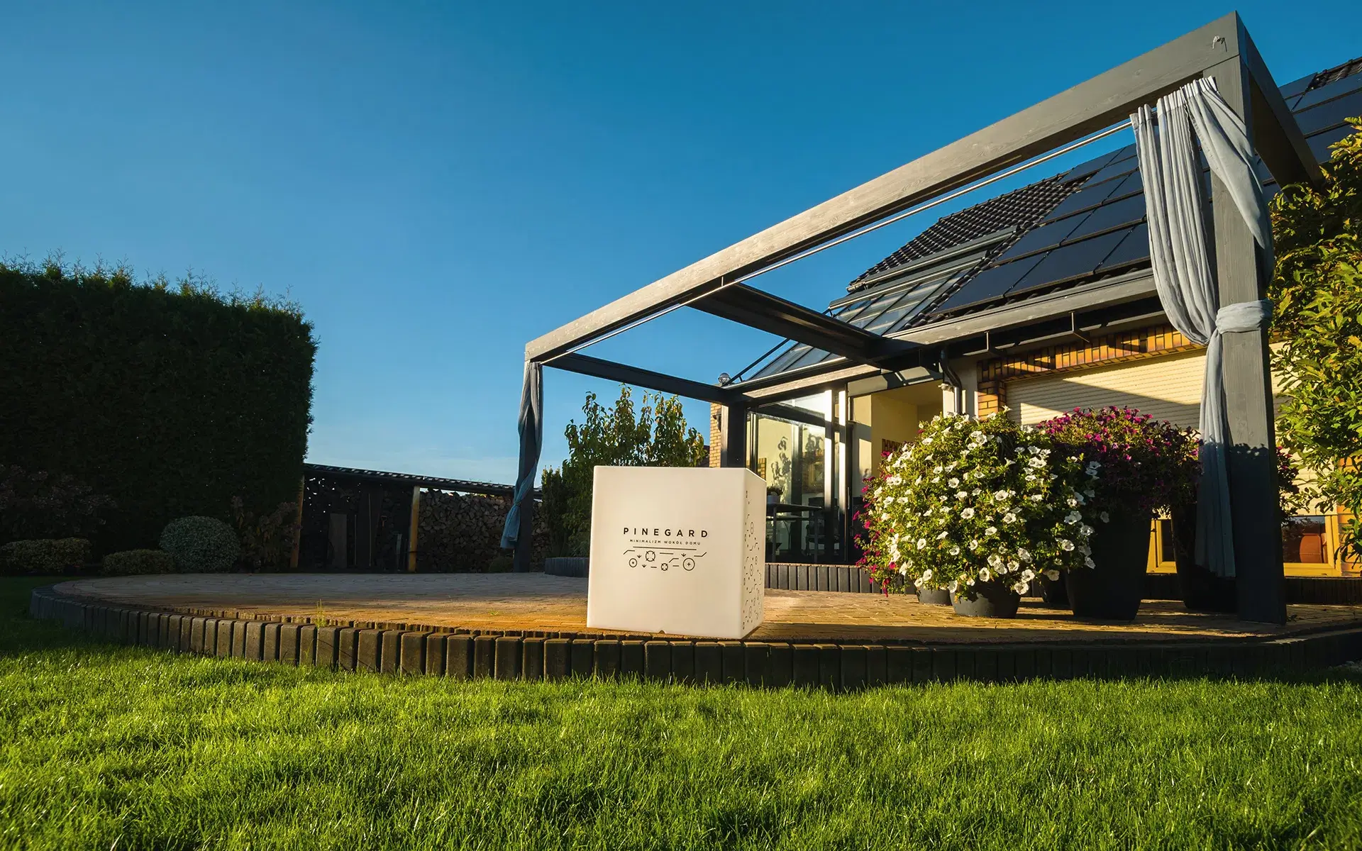 Auf dem Bild ist eine moderne Pergola mit schlankem Rahmen neben einem Haus mit Solarpanelen auf dem Dach zu sehen. Auf der Holzterrasse stehen große Blumentöpfe mit blühenden Pflanzen und ein dekoratives Element mit dem Pinegard-Logo.