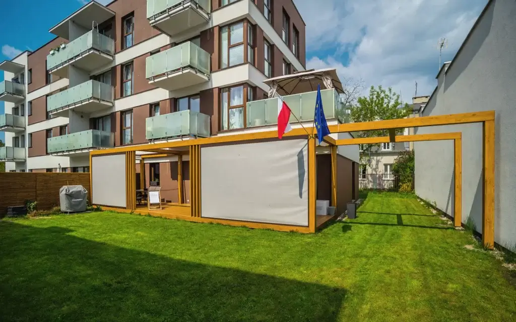 Ich sehe ein modernes Wohngebäude mit mehreren Balkonen. Davor gibt es einen Garten mit einer überdachten Terrasse und zwei Fahnen.