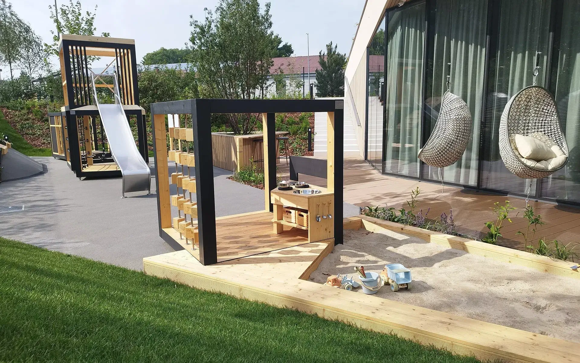 Die Grafik zeigt einen modernen Spielplatz im Freien. Im Vordergrund befindet sich ein Sandkasten mit Spielzeug und eine hölzerne Spielküche. Daneben hängen zwei Korbsessel mit Kissen. Im Hintergrund ist eine größere Kletter- und Rutschstruktur zu sehen. Die gesamte Anlage ist von Grün umgeben – Gras, Bäumen und Sträuchern.