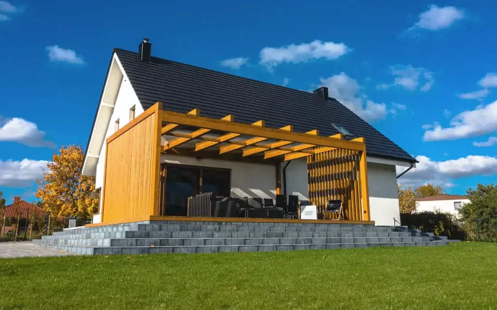 Das Foto zeigt ein modernes Haus mit einem schwarzen Satteldach und einer weißen Fassade, daneben eine geräumige Terrasse mit einer Holzpergola. Die Konstruktion der Pergola ist mit horizontalen Balken und vertikalen Lamellen ausgestattet, die teilweisen Schatten und Privatsphäre bieten. Auf der erhöhten Terrasse, die über breite Treppen aus grauen Platten zugänglich ist, steht eine Gartenmöbelgarnitur. Die Umgebung wird durch einen gepflegten Rasen, Bäume in herbstlichen Farben und die im Hintergrund sichtbaren Nachbarhäuser geprägt.