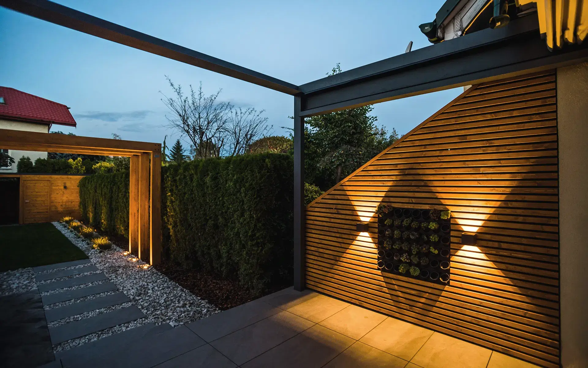 Auf dem Bild sieht man einen modernen Gartenbereich mit einer Holz- und Metallpergola bei Abendbeleuchtung. An der Holzwand befindet sich eine dekorative Pflanzeninstallation, daneben sieht man einen Weg mit Steinplatten, weißen Kies und hohe Hecken.