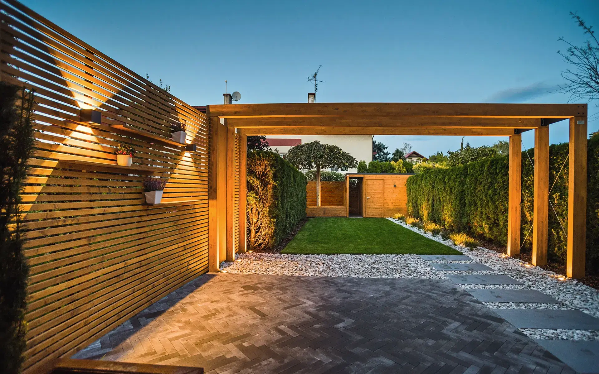 Auf dem Bild sieht man einen modernen Garten mit einer großen Holzpergola und Beleuchtung am Abend. Es gibt einen gepflasterten Bereich, weißen Kies, einen Rasen, Hecken und eine Holzhütte im Hintergrund.