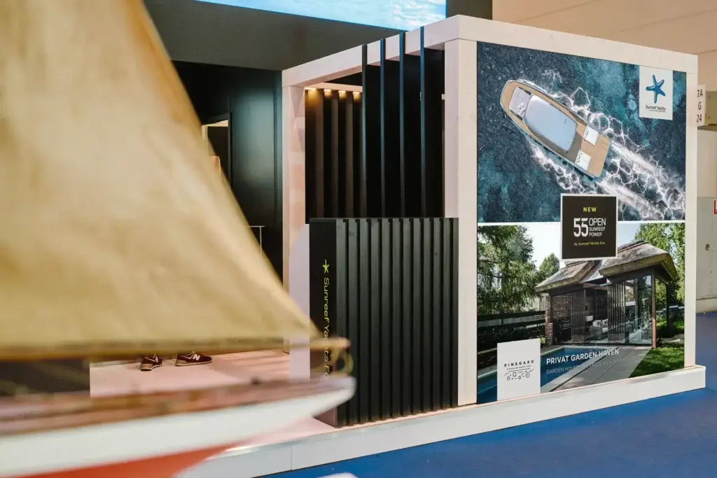 Na zdjęciu widać nowoczesne stoisko targowe firmy Sunreef Yachts Eco w hali wystawowej. Na jednej ze ścian umieszczono duże grafiki przedstawiające jacht z lotu ptaka oraz wizualizację ogrodowego pawilonu, wraz z informacją o modelu „55 Open Sunreef Power”.