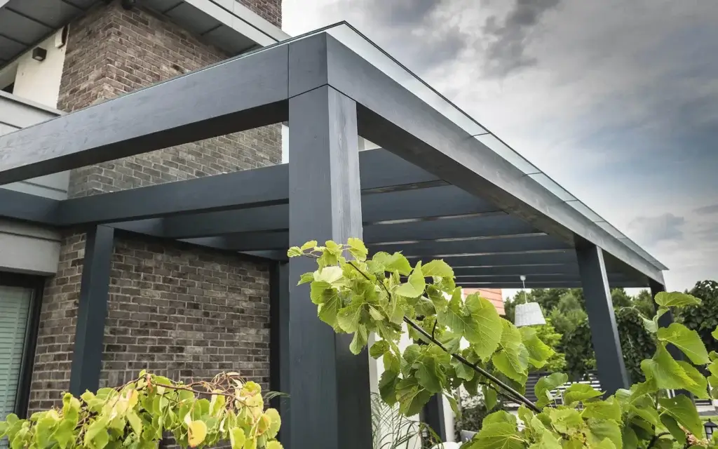 Nowoczesna pergola przy ceglanej elewacji — ciemne, metalowe belki tworzą geometryczną strukturę, zapewniając półcień i elegancki kontrast z naturalną zielenią. Minimalistyczna forma, stylowe połączenie architektury z otoczeniem.
