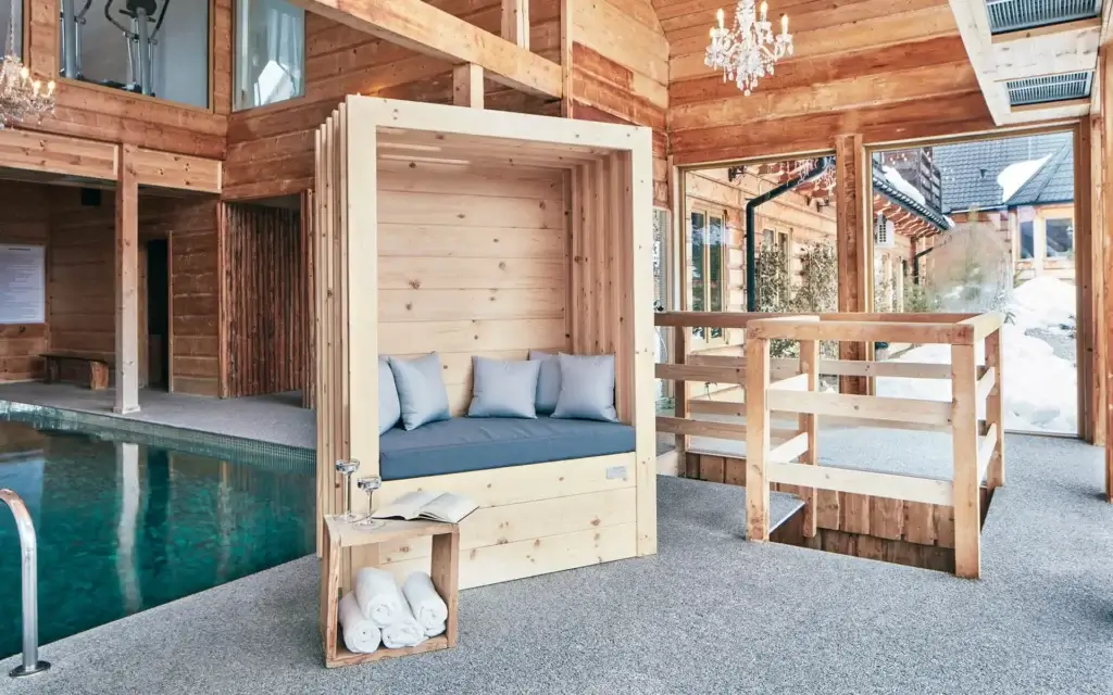 Luksusowa strefa wellness z drewnianą architekturą i przeszklonymi ścianami — kameralny basen, designerska kapsuła wypoczynkowa z poduszkami, miękkie światło i naturalne materiały tworzą atmosferę spokoju i elegancji. Idealne miejsce na regenerację w stylowym otoczeniu.