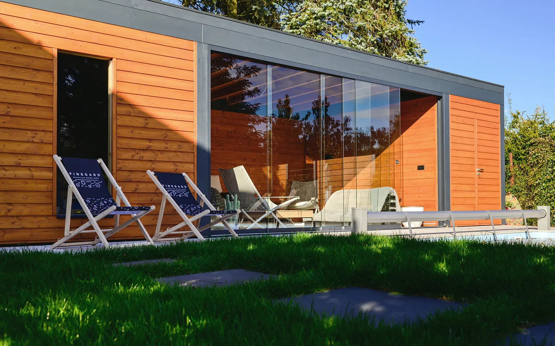 Das Foto zeigt ein modernes Gartenhaus mit einer Fassade aus horizontalen Holzpaneelen in natürlichem Farbton, umrahmt von dunkelgrauen Leisten. Große Verglasungen geben den Blick auf das minimalistische Interieur mit Sesseln und einem Tisch frei. Auf dem grünen Rasen vor dem Gebäude stehen zwei blaue Liegestühle mit weißen Gestellen und der Aufschrift „PINEGARD“. Der Hintergrund wird von Bäumen und Hecken geprägt, was dem Raum einen ruhigen, intimen Charakter verleiht.