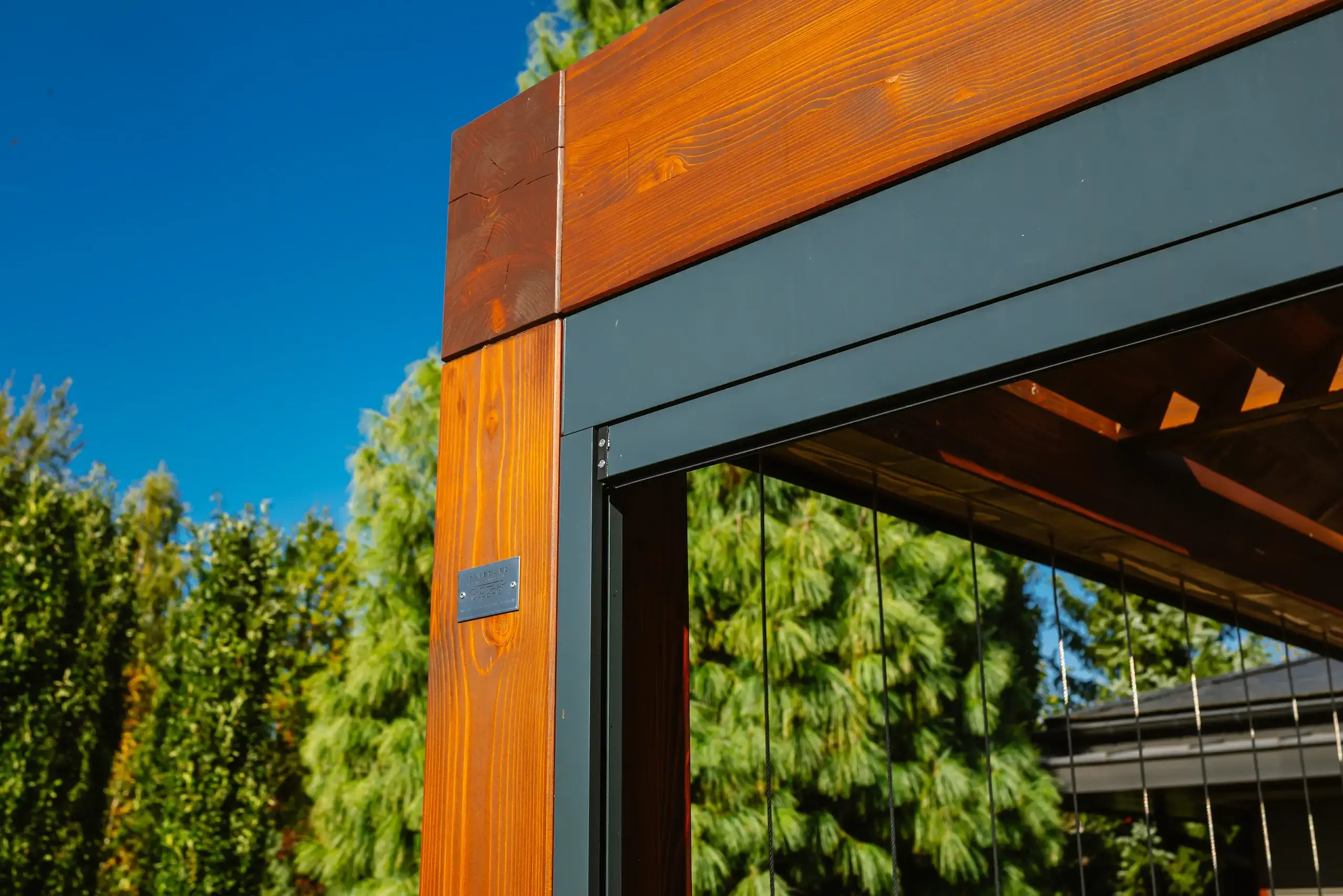 Auf dem Bild sieht man die Ecke einer modernen Holzpergola mit einer stabilen Konstruktion. Die Holzbalken haben eine warme braune Farbe und sind mit dunklen Metallelementen verbunden. Im Hintergrund erkennt man grüne Bäume und einen klaren blauen Himmel.